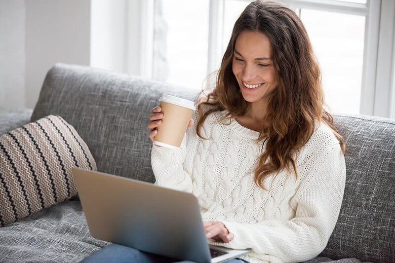 Woman Using Her Laptop Computer While Drinking Coffee on Her Couch